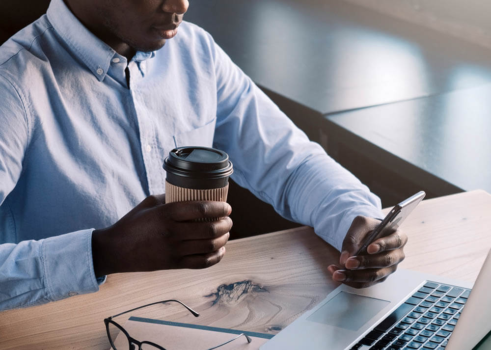 man working on his laptop while holding phone and coffee